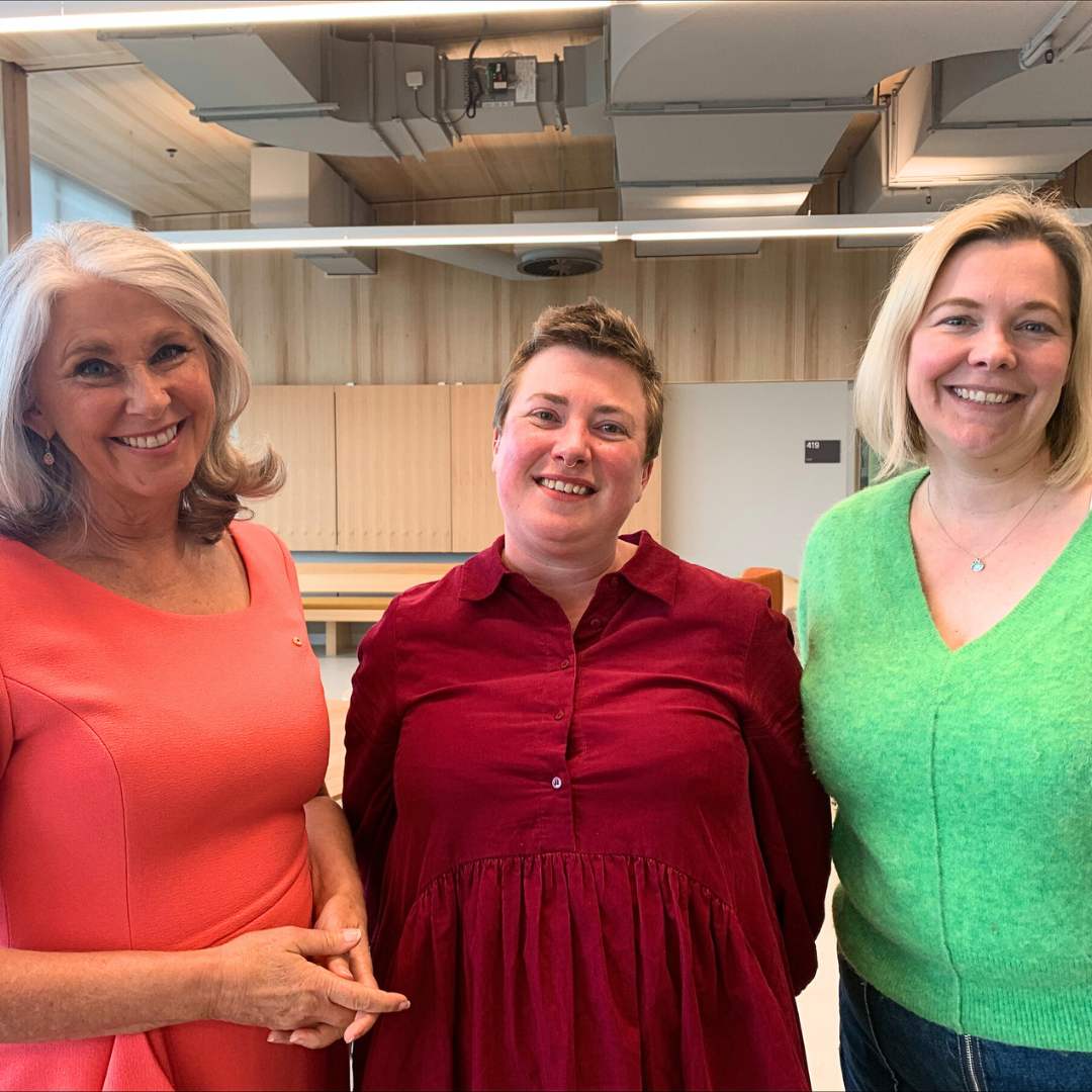 Photo of Tracey Spicer smiling in a red dress, standing with two other smiling white women, one in a red shirt and one in a bright green jumper.