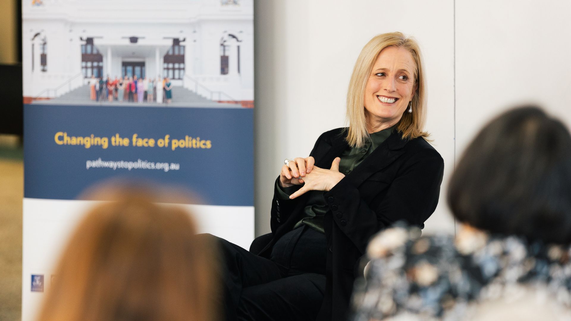 Senator Katy Gallegher, a white woman with long blond hair, wearing black and holding and looking off to the side, smiling. There is a Pathways to Politics banner in the background.