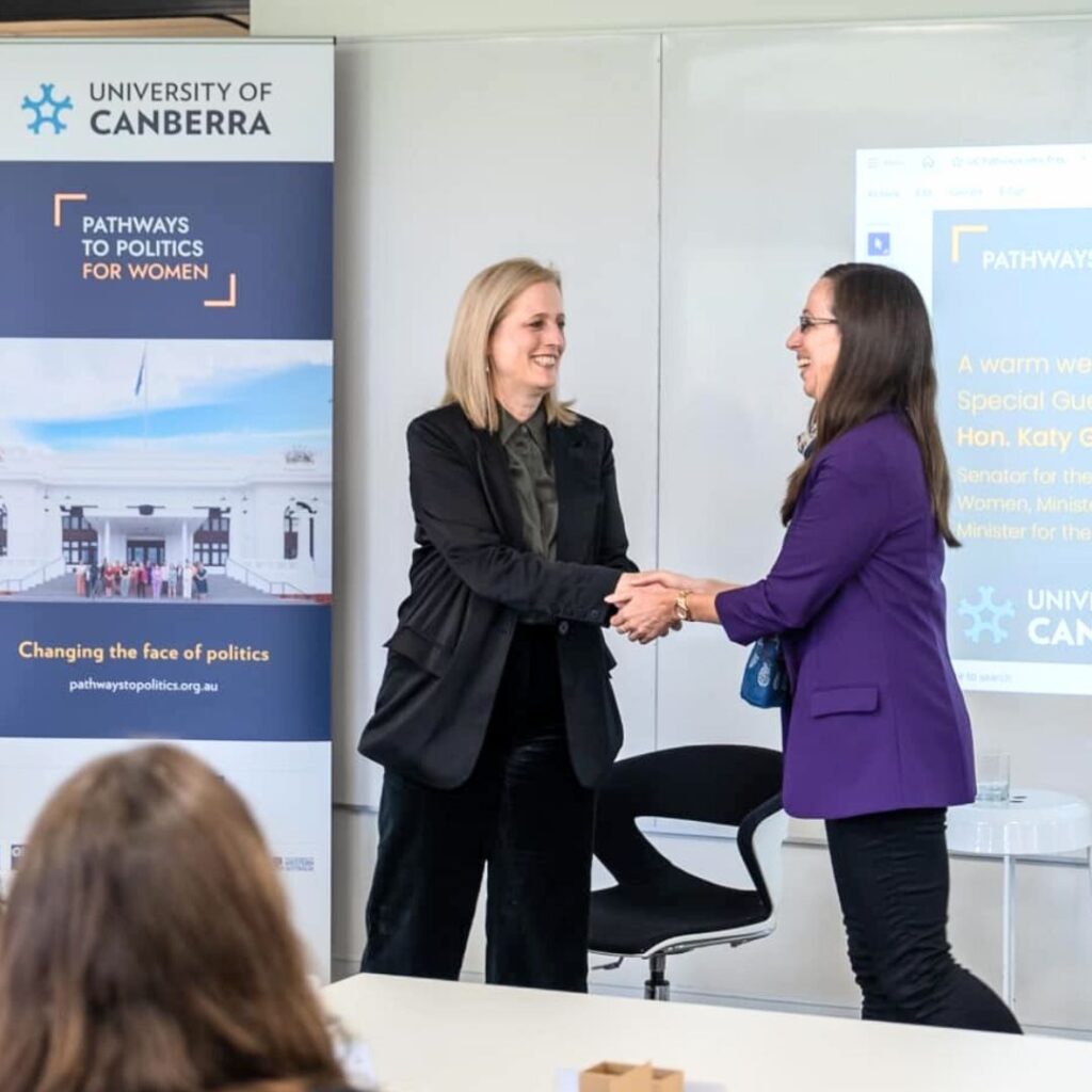 Photo of Dr Leonora Risse shaking hands with Senator Katy Gallagher in front of a Pathways to Politics banner