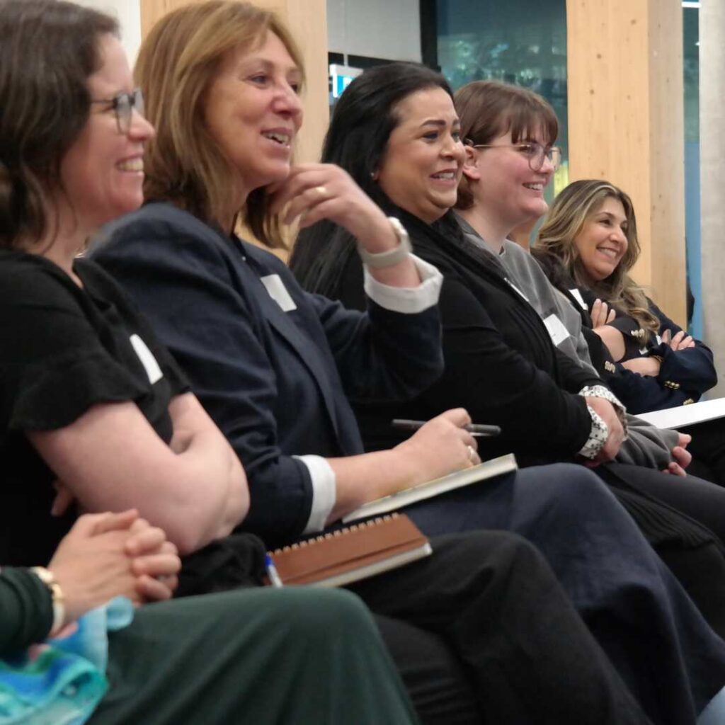 Five diverse women seated in an audience. They have notebooks in their laps and are smiling.