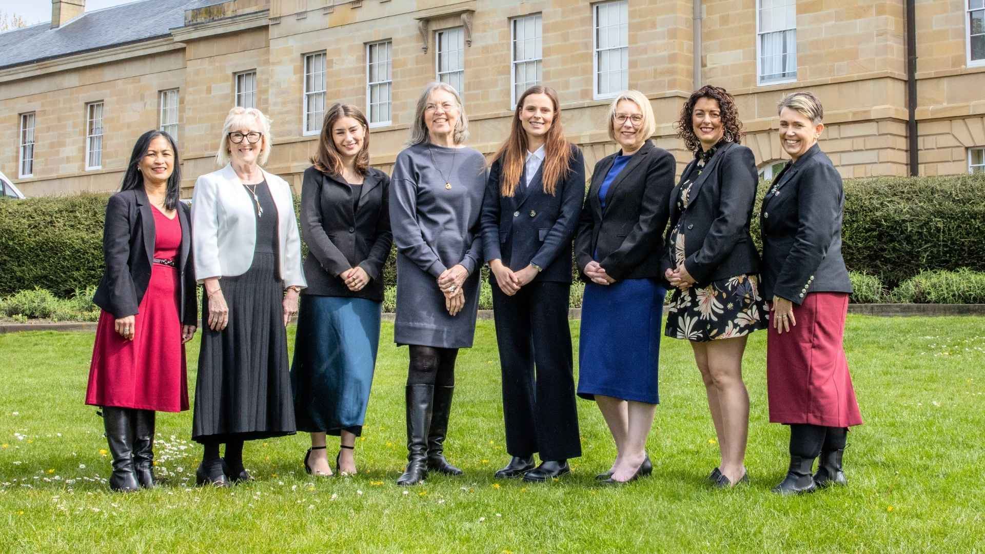 Eight diverse women standing on a green lawn in front of sandstone parliamentary building