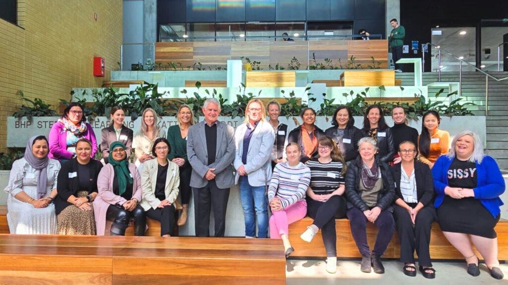 WA cohort, a group of more than 20 diverse women, photographed in a group in a modern learning space. Ken Wyatt is standing in the centre of the group.