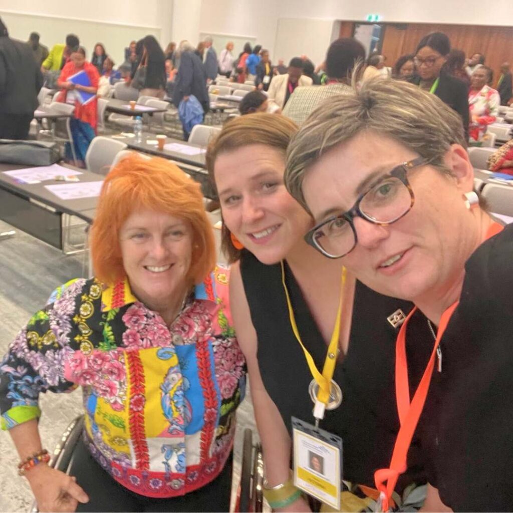 Selfie of three women, two have lanyards around their necks. In the background there is conference style seating and a crowd of women talking.