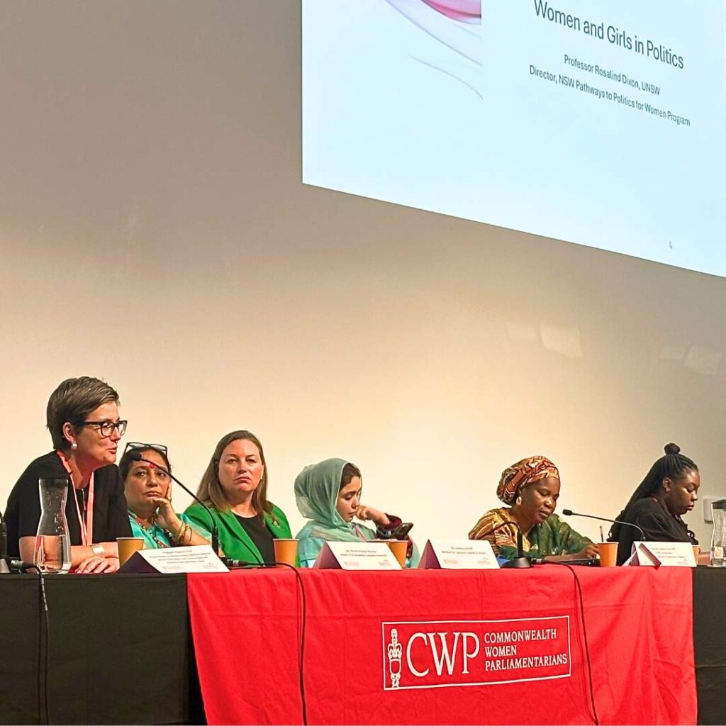 Six diverse women sitting behind a table on stage, speaking into microphones