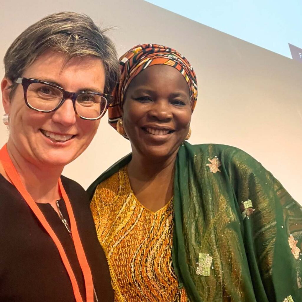 Selfie of a white woman with short hair and glasses, orange lanyard around her neck, and a woman of African appearance with a colourful headscarf and green and yellow blouse