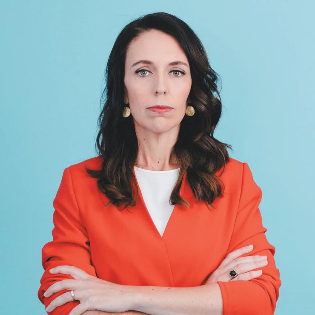 Photo of Jacinda Ardern wearing a tangerine coloured blouse, arms crossed and serious expression