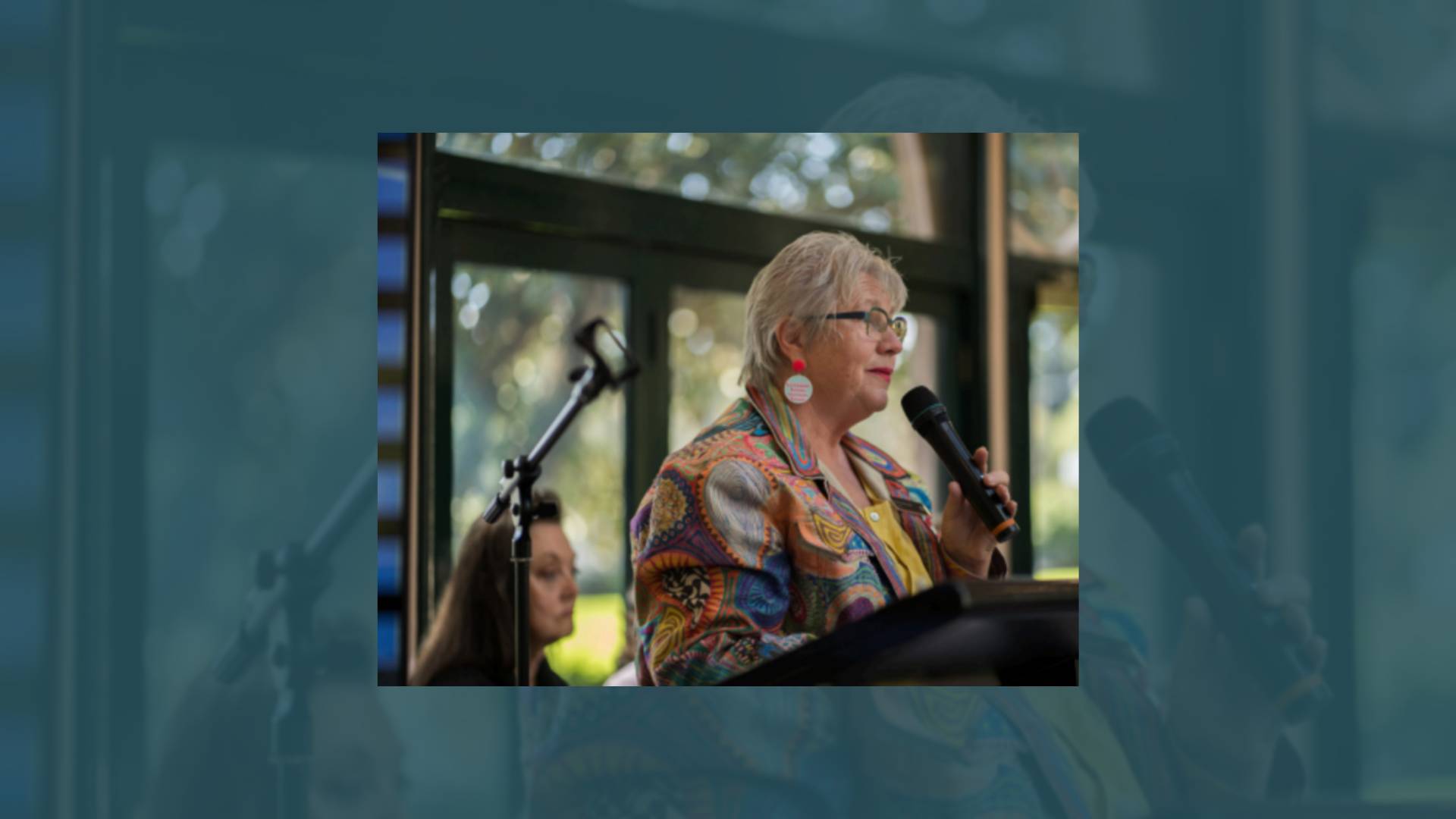 White woman wearing glasses and a colourful blouse is standing at a podium holding a microphone