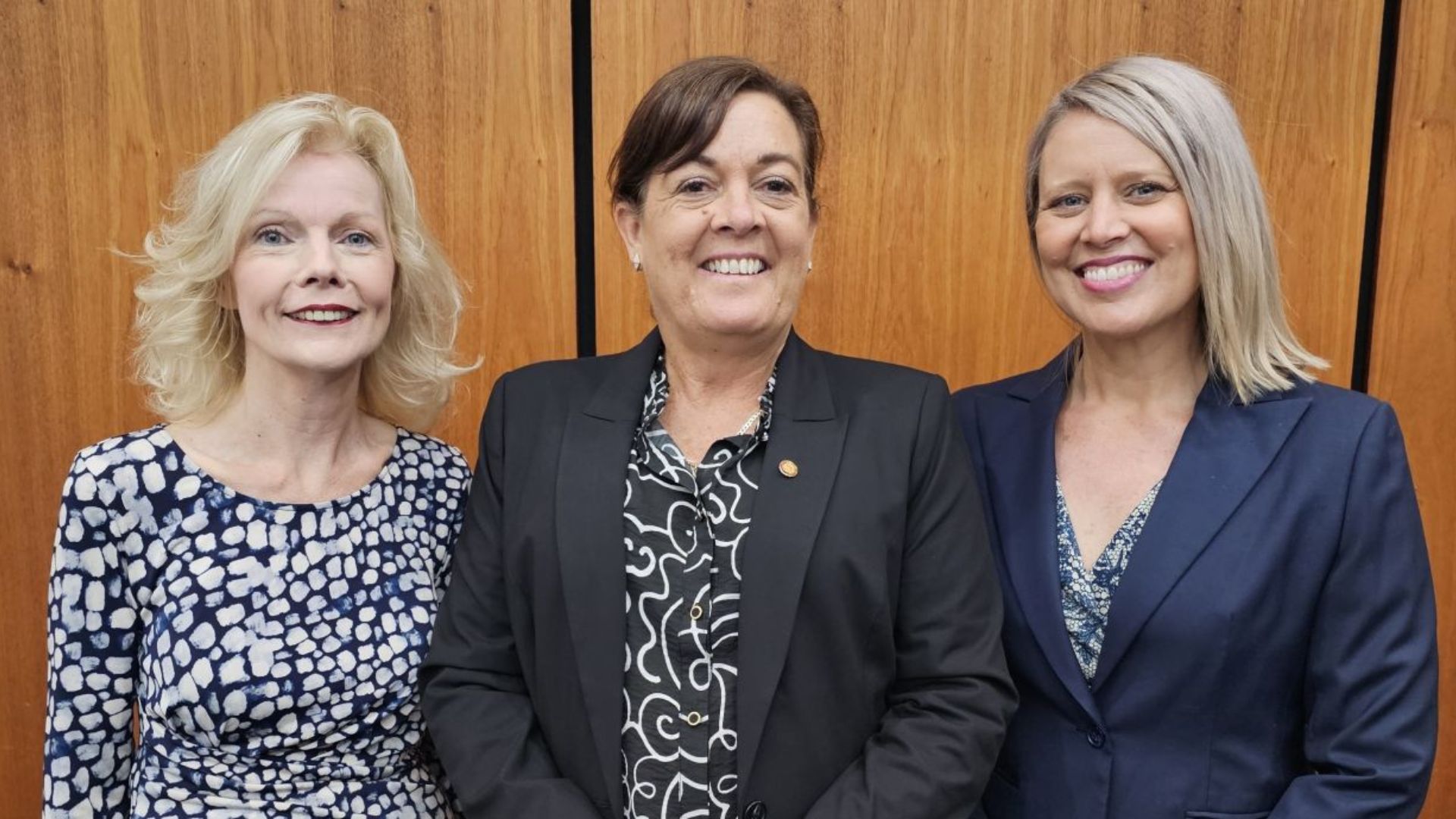 Three professional women pose smiling for a photo agains wood panel background