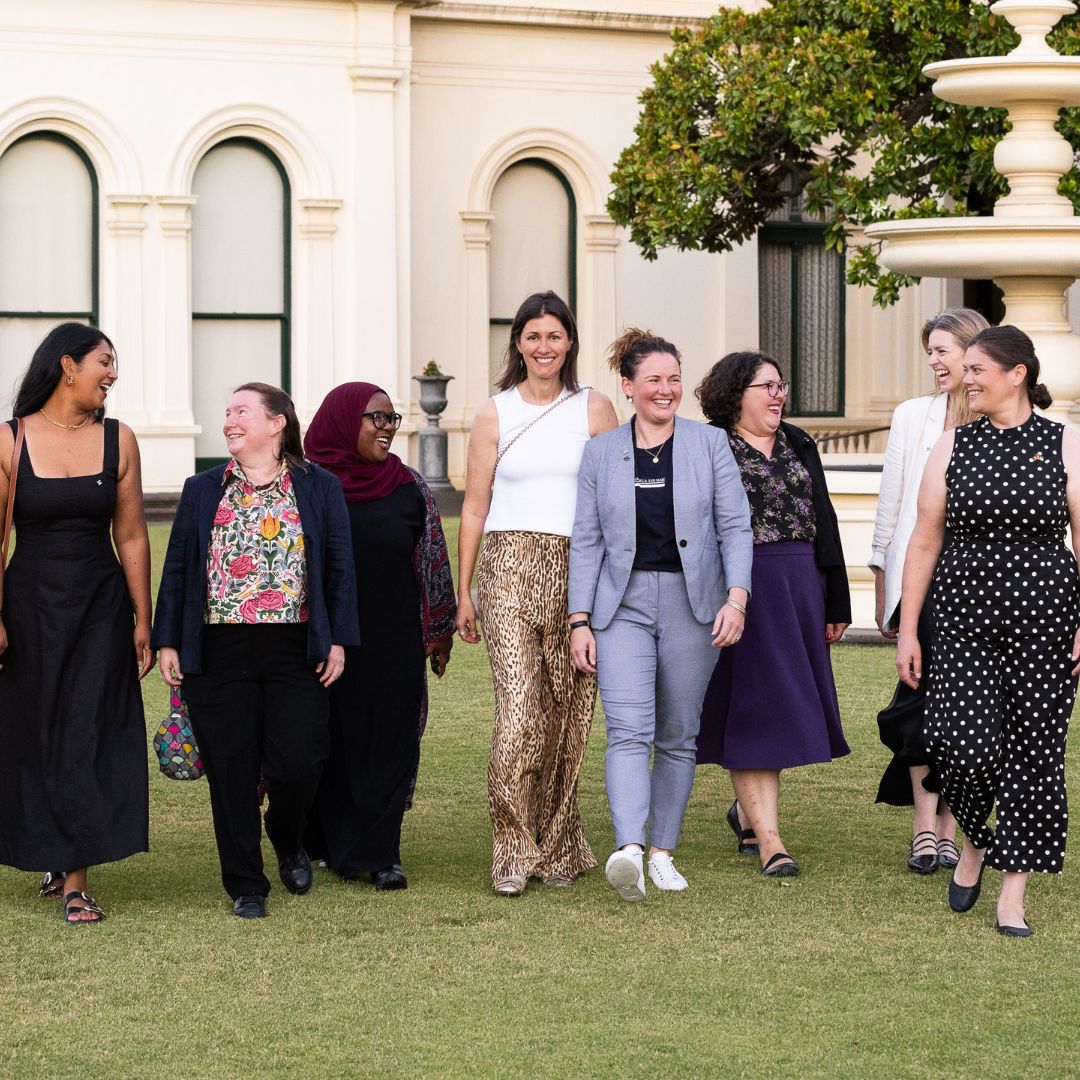 Photo of eight diverse women walking on a green lawn outside grand building. They are smiling. Some are wearing suits, others long dresses.