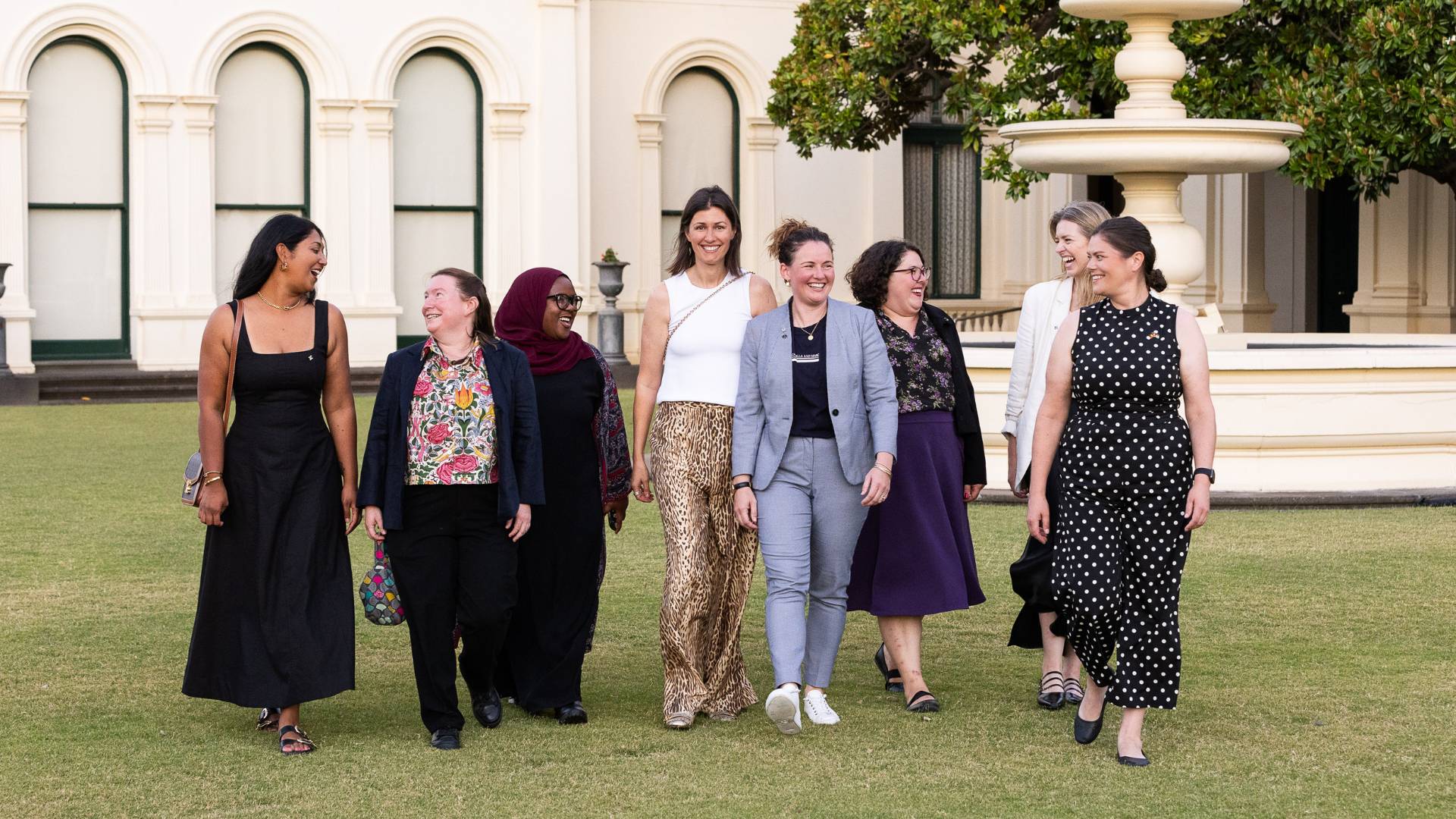 Photo of eight diverse women walking on a green lawn outside grand building. They are smiling. Some are wearing suits, others long dresses.