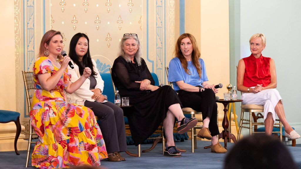 5 women sit on a stage in a panel discussion. One in a bright yellow and red dress is holding a microphone