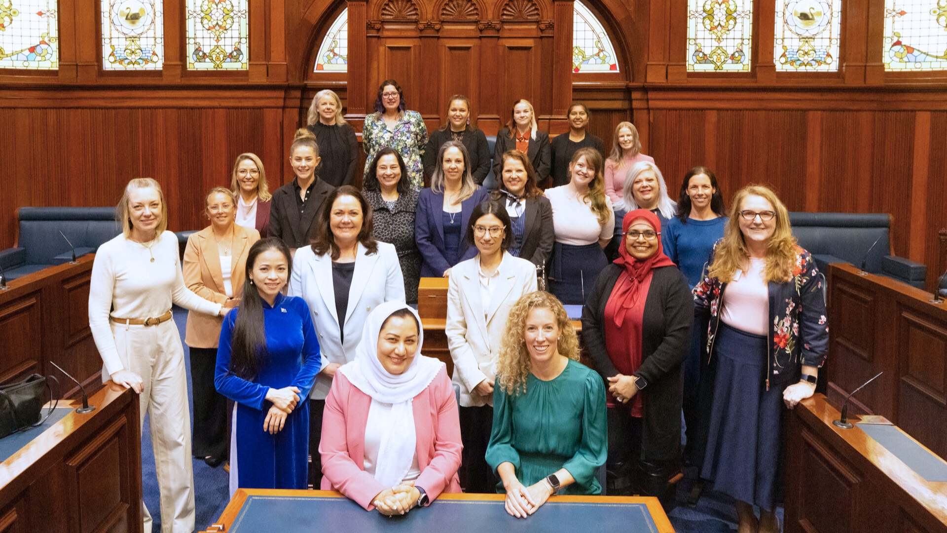 Group of diverse women standing in 4 rows inside a wood paneled parliament chamber