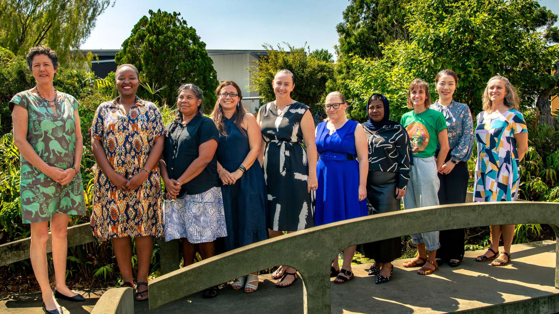 Group of 10 diverse women wearing summery brightly coloured dresses and skirs pose for a photo on a bridge, There is lots of greenery behind them.