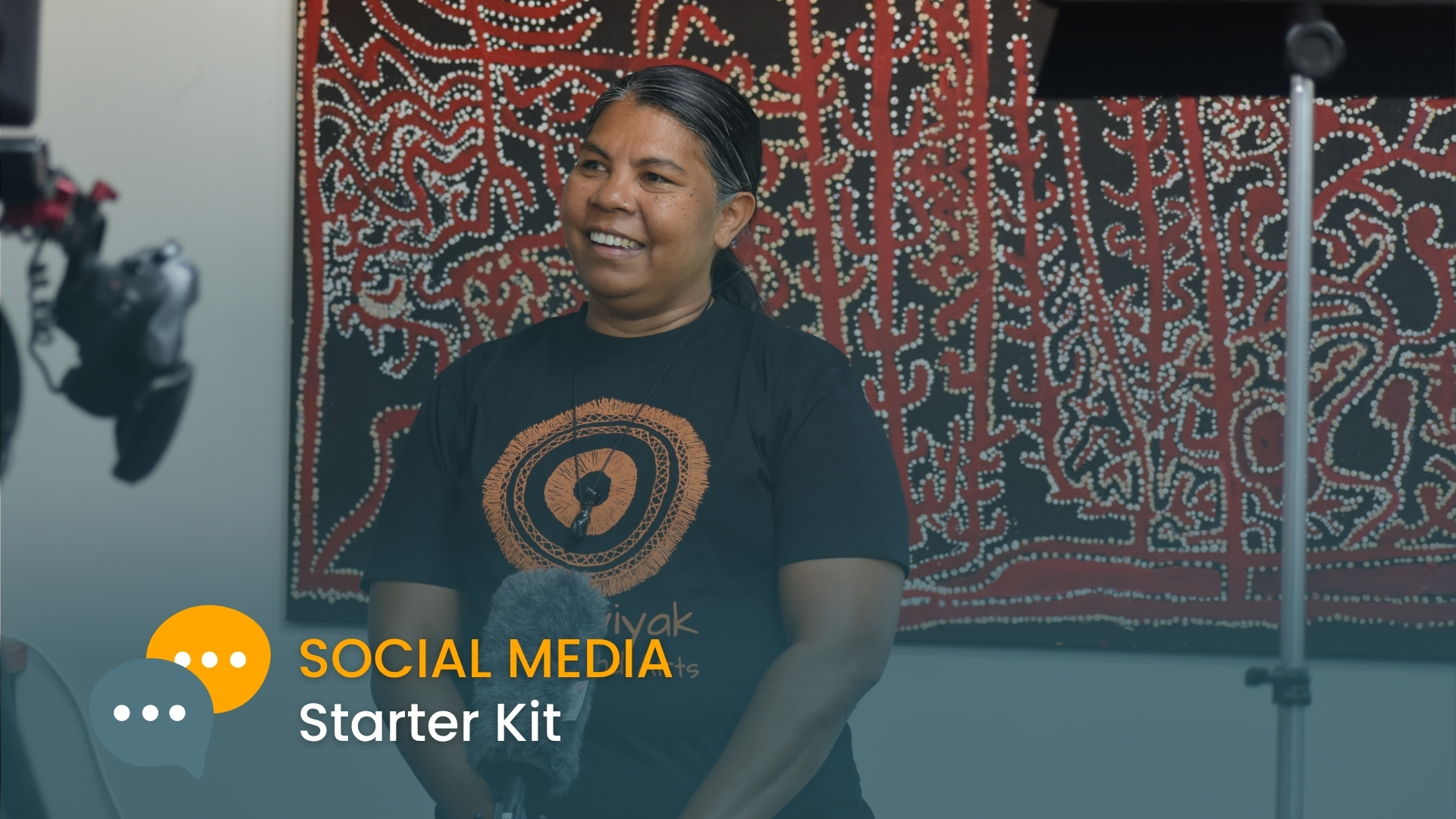 Photo of a smiling woman being interviewed with microphone, against a backdrop of larce Aboriginal artwork