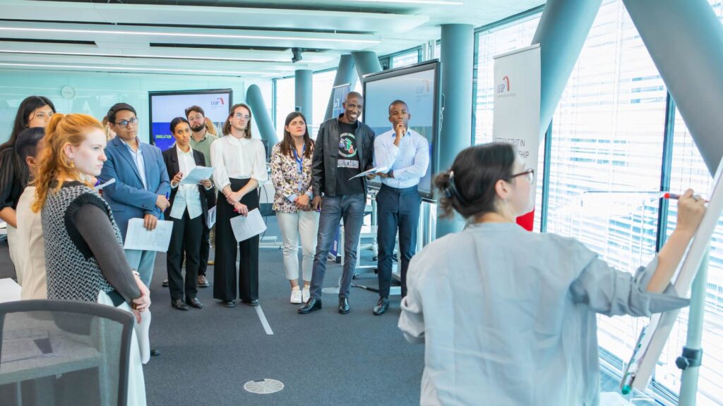 Group of diverse young people stand in a semicircle in a bright, modern conference space. A woman in the foreground is writing on a whiteboard.