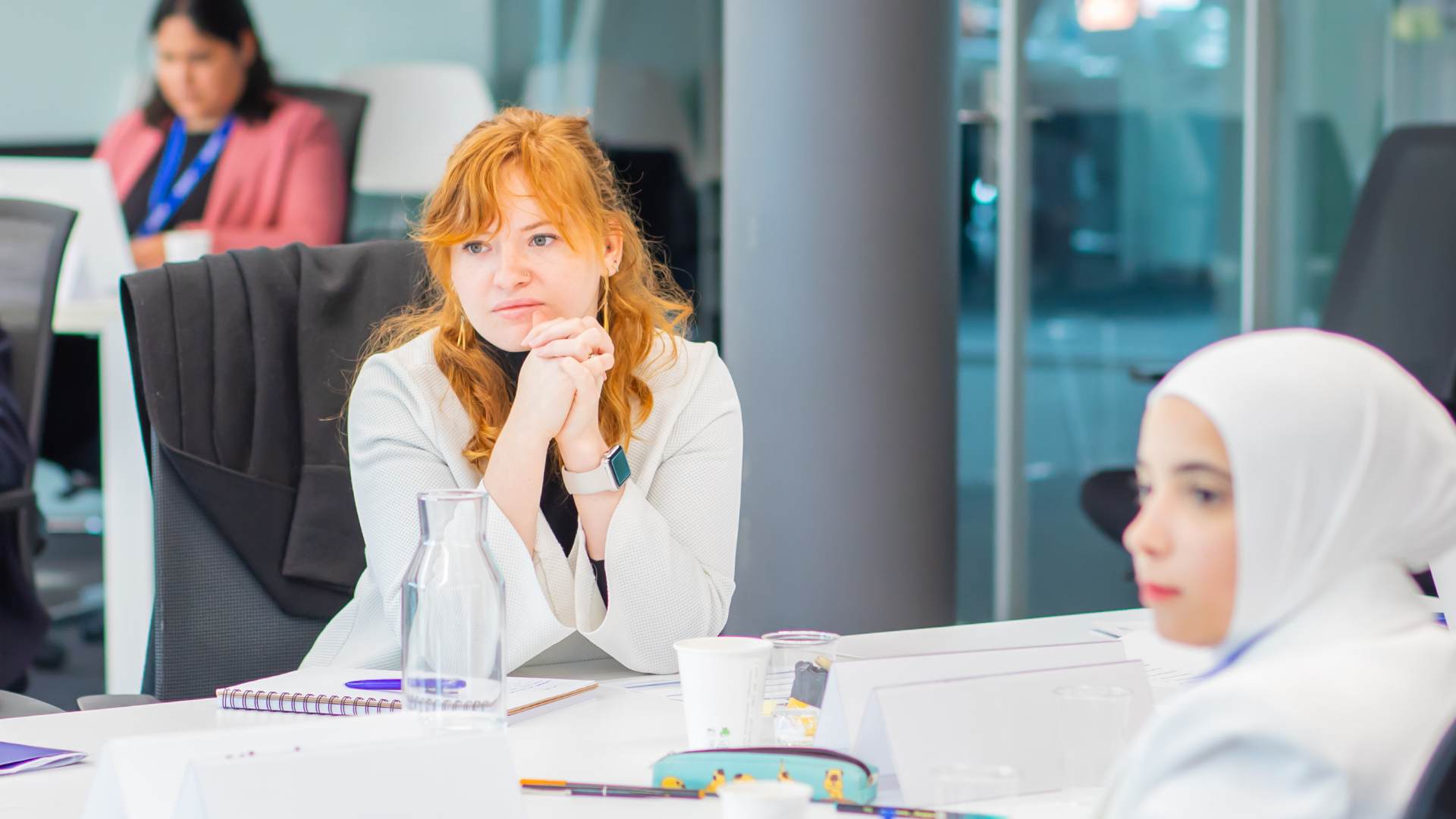 Photo of Neane Carter, leaning on her hands in a modern conference room setting, looking into the distance. She has long auburn hair and is wearing a white blouse. There is a young woman wearing a hijab in the foreground, and a middle aged woman in pink blazer in the background.