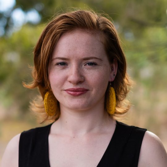 Smiling portrait photo of Nenae Carter. She has auburn hair and is wearing a short sleeved black top, trees in the background