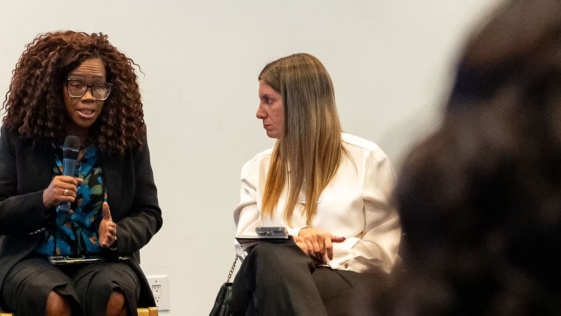 Woman with dark skin and curly hair, wearing glasses, is sitting talking into a microphone. A white woman with long hair sits next to her listening seriously.