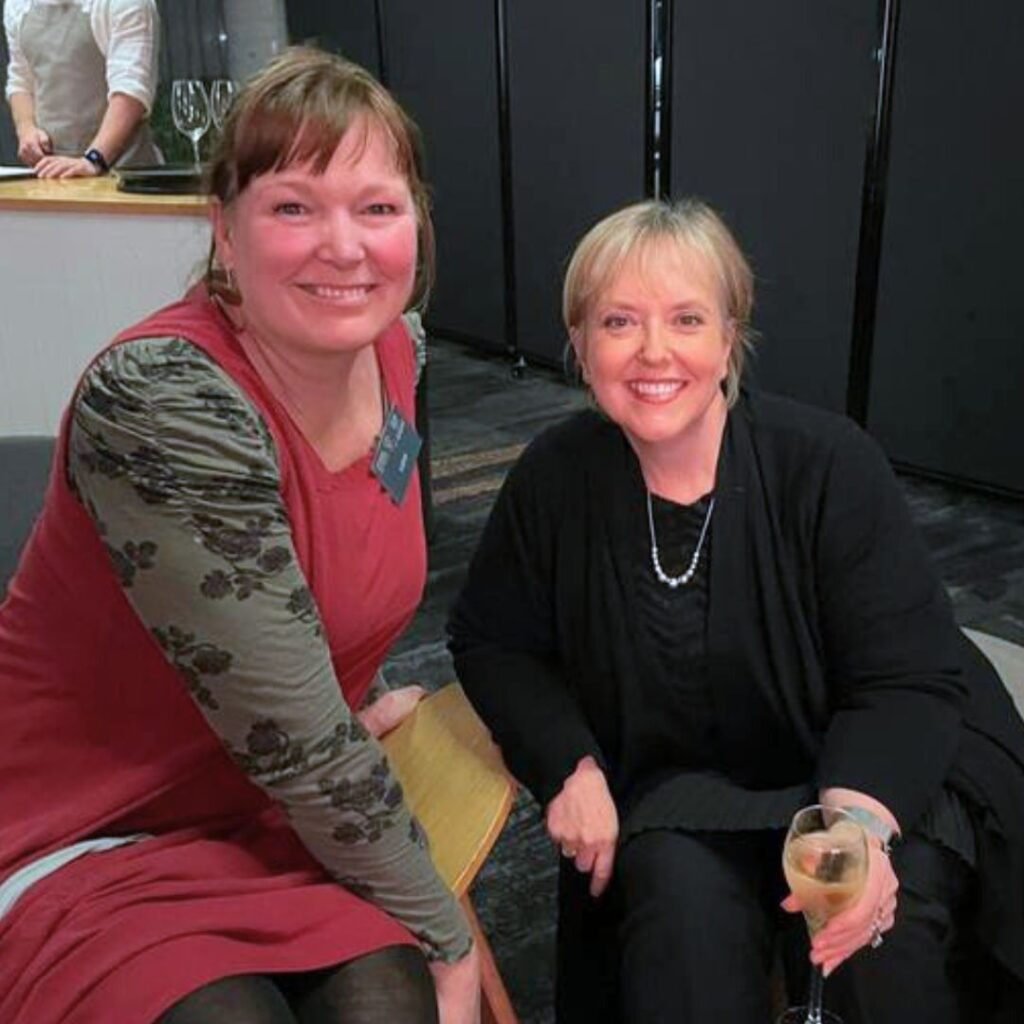 two white women sit smiling and posing for a photo. One is holding a glass of champagne.