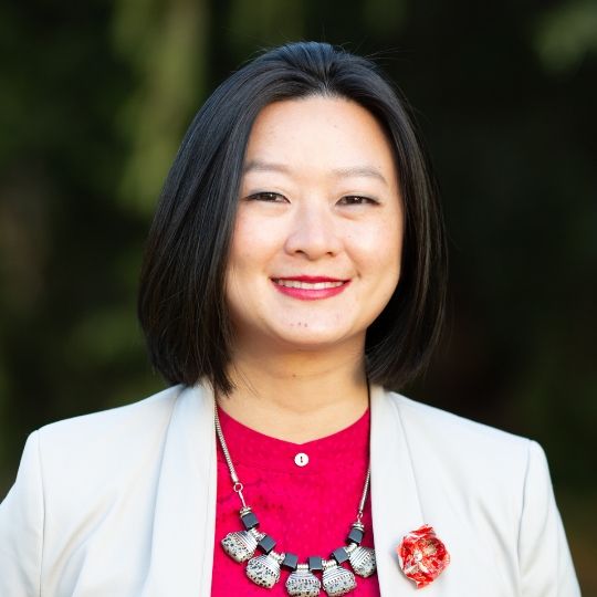Photo of a smiling asian woman with bobbed hair, wearing a cream blazer over a bright pink blouse