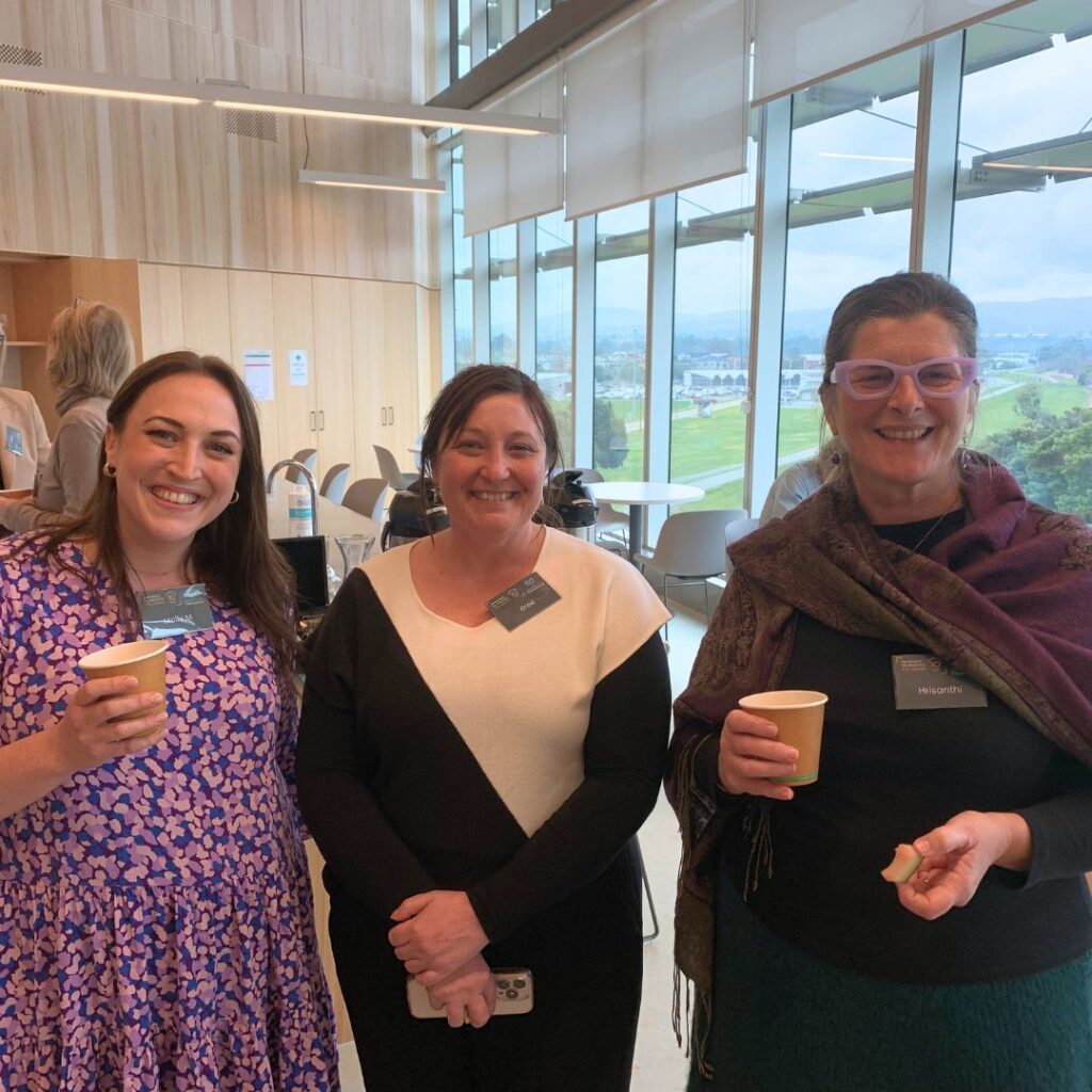 three diverse women holding coffee cups, smiling and posing for the photo. Green hills can be seen outside the window behind them.