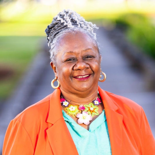 Photo of a smiling woman, dark skinned wearing bright orange blazer , green blouse and large colourful necklace
