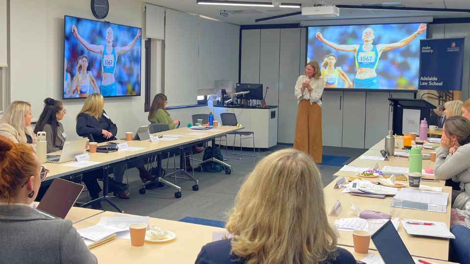 A woman with long blond hair presenting to a group of women seated at U-shaped tables
