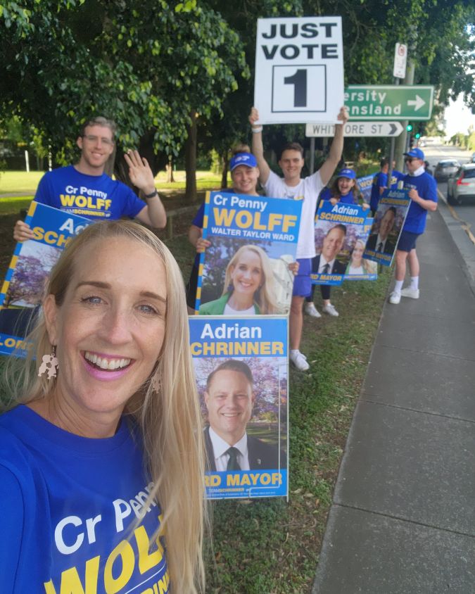 Selfie of a woman with long blonde hair wearing a blue campaign tshirt, crowd of people in the same tshirts holding "Penny Wolff" political placards behind her