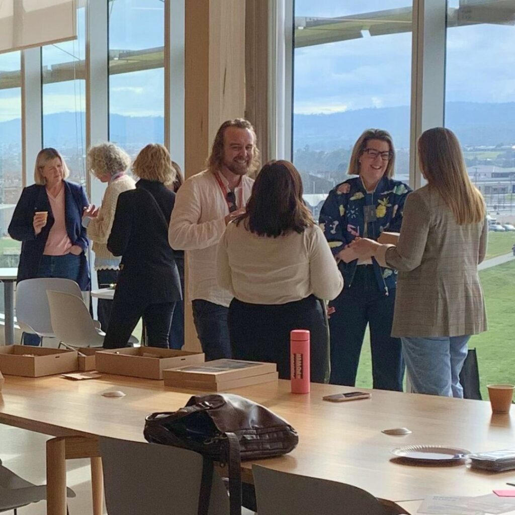 Six women and one man chatting and drinking coffee, standing in a classroom space in front of large windows with green hills outside