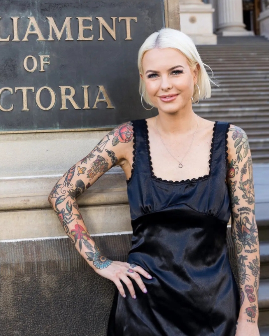 Photo of a woman wearing a sleeveless black dress outside parliament, with two full sleeves of tatoos.