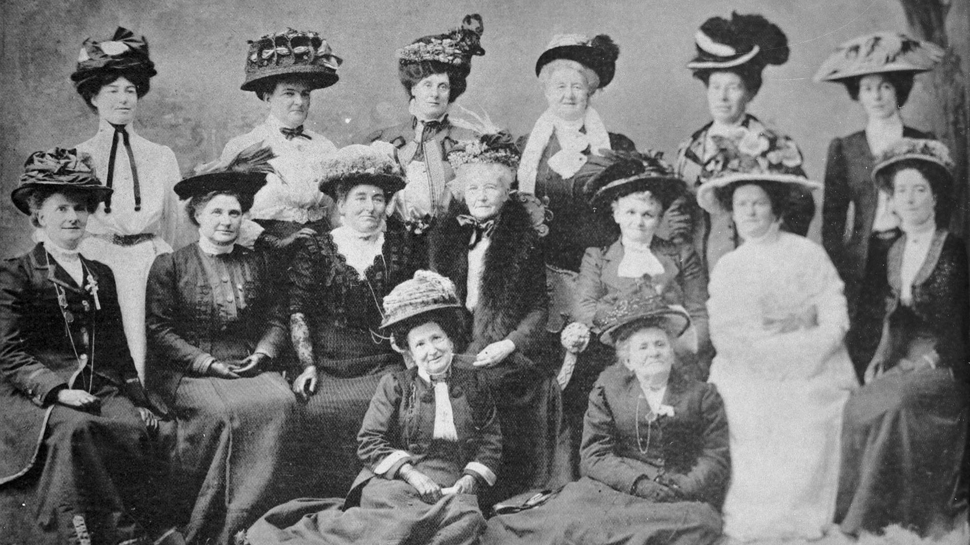 Victorian era black and white photograph of 15 white women in long dresses and large decorated hats, posed in three rows