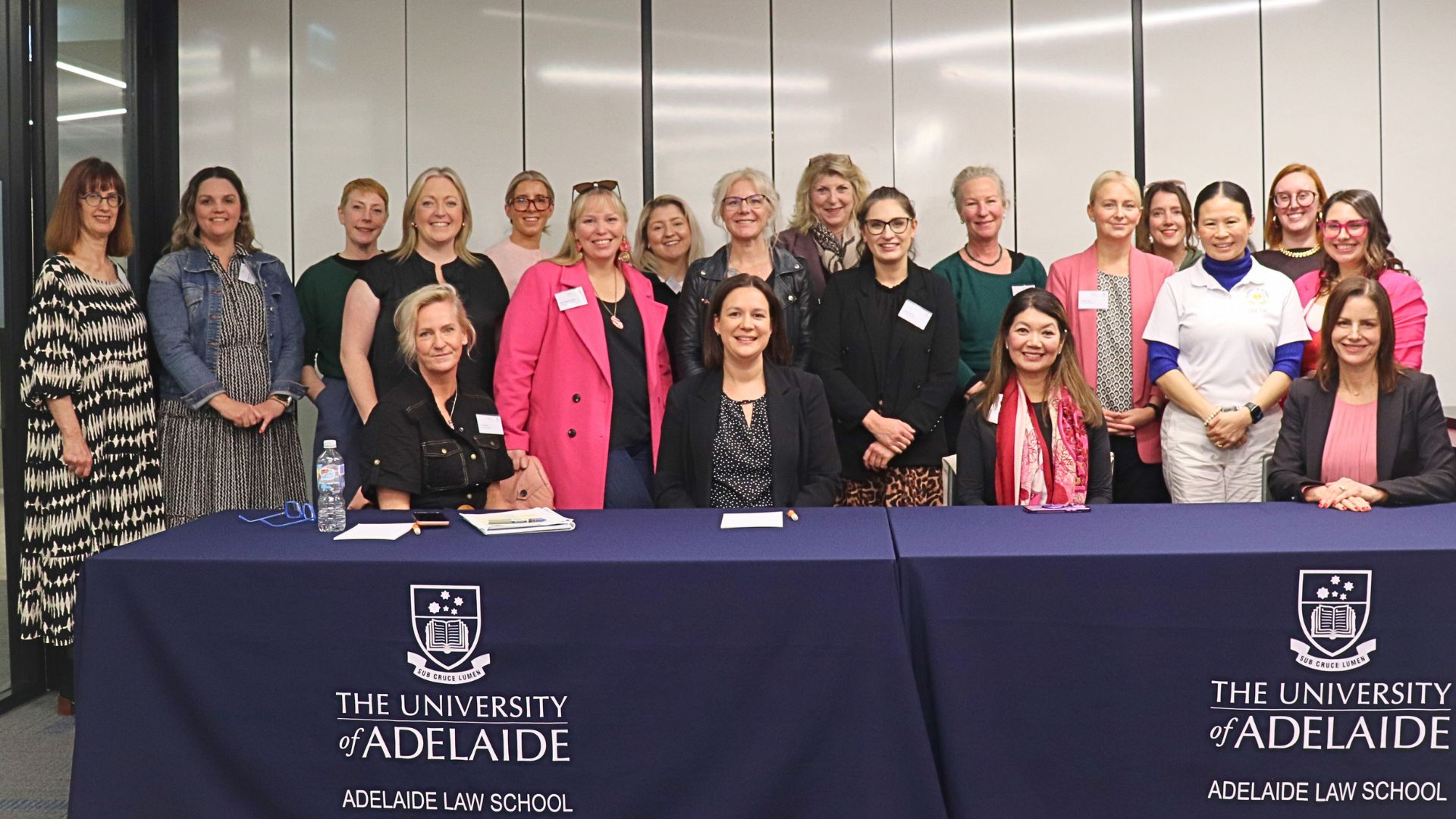 Large group of diverse women stand behind low tables draped with blue cloth, the University of Adelaide logo printed on them