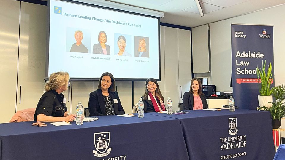 Four women on a panel, smiling and talking seated in front of a projector screen