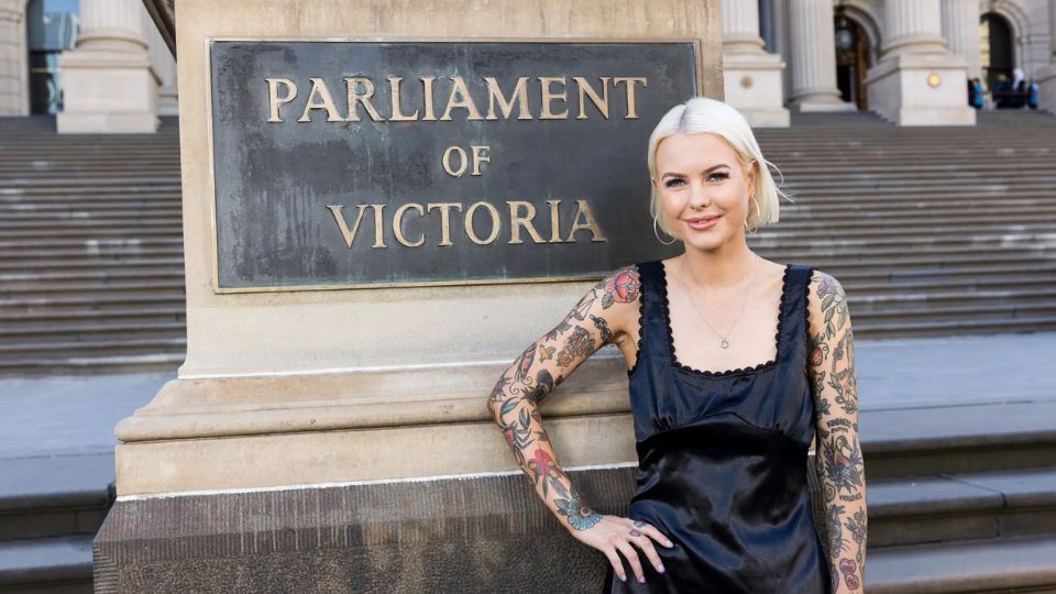 Photo of Georgie Purcell, a young blonde woman with tattooed arms, smiling next to a sandstone pillar with a sigh "Parliament of Victoria" on it