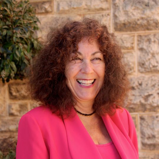 Smiling photo of Hilary Silbert, curly red-brown hair, wearing a bright pink blazer.