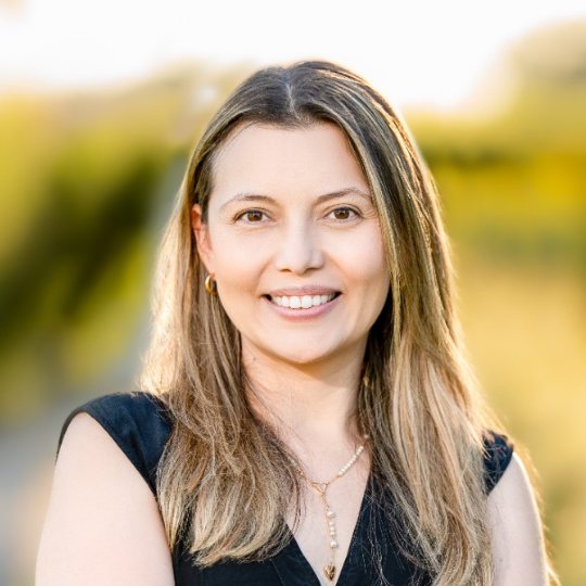 Councillor Ivonne Rodriguez smiling, with long light brown hair, wearing a short-sleeved black top. Trees are blurred in the background.
