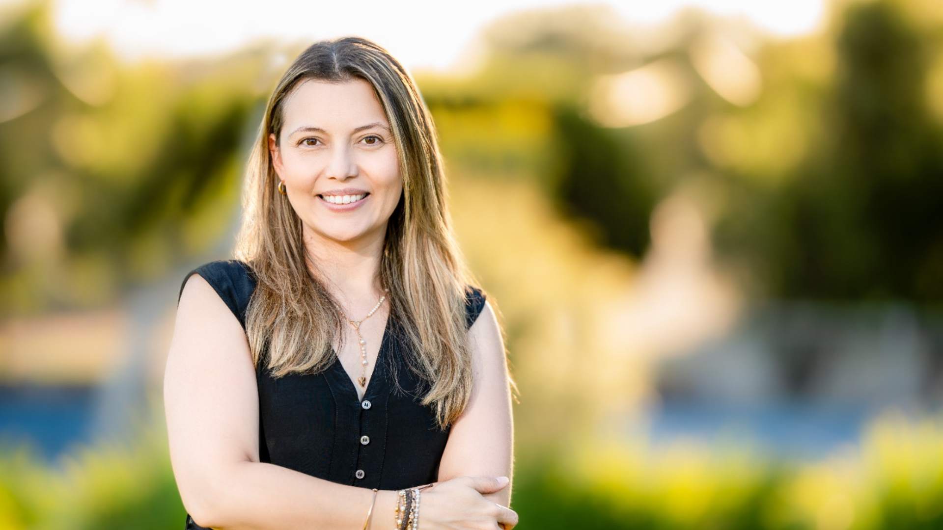 Councillor Ivonne Rodriguez smiling, with long light brown hair, wearing a short-sleeved black top. Trees are blurred in the background.