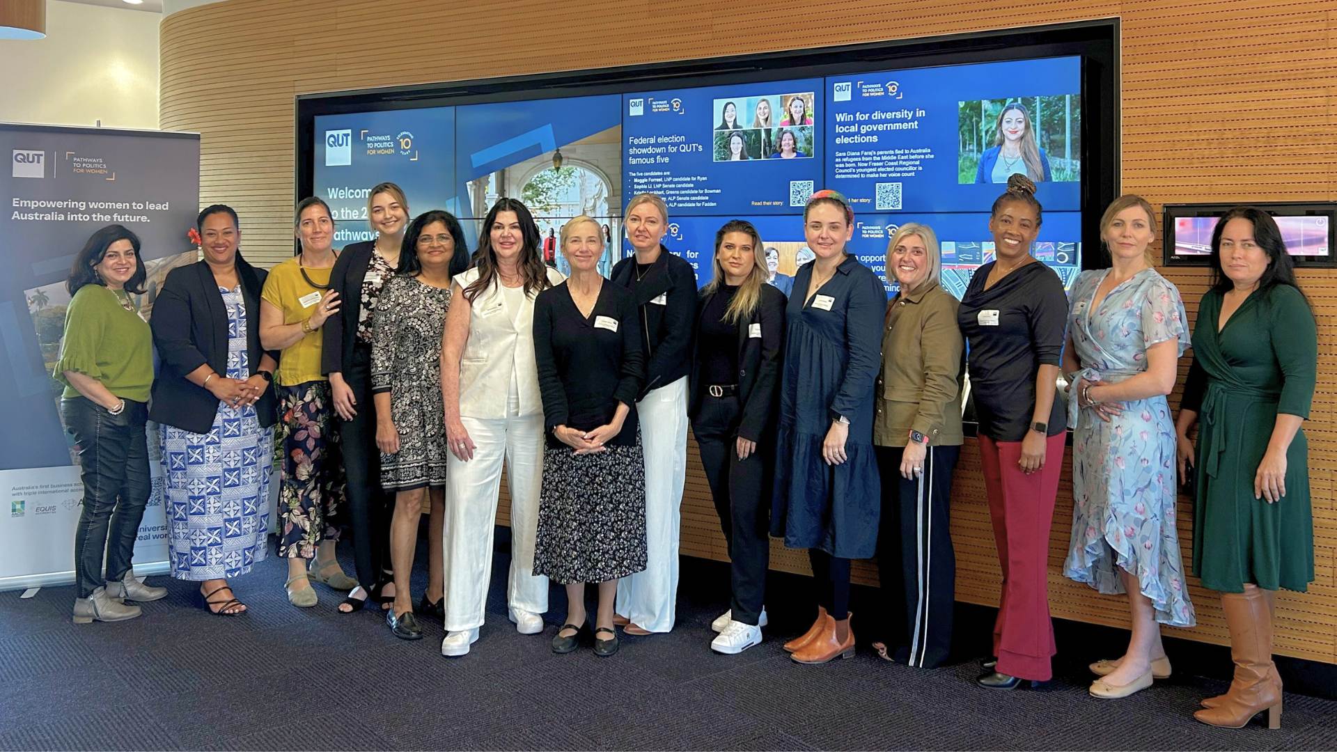 Large diverse group of women pose smiling in front of blue media screens in a large modern room