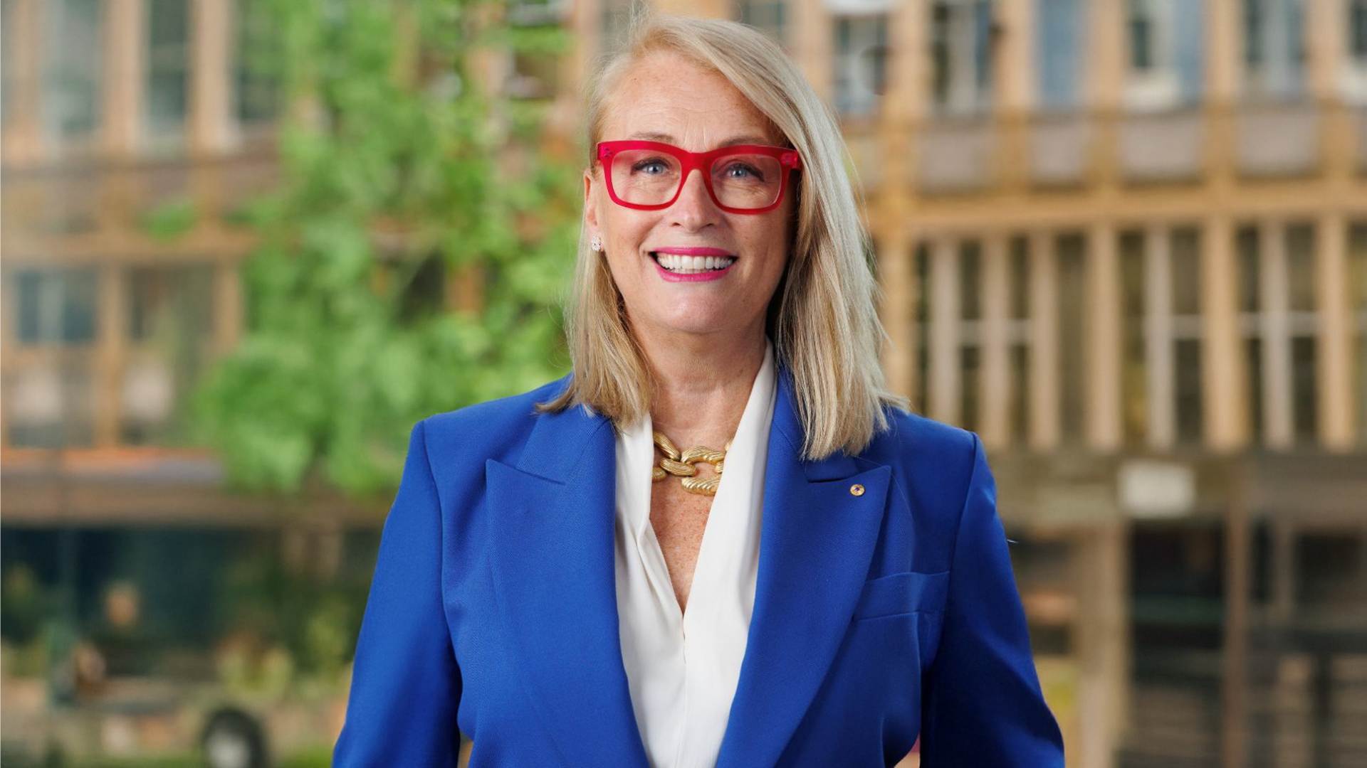 Photo of Sally Capp smiling, old building and greenery in the background. Sally is wearing a bright blue blazer and bright pink glasses.