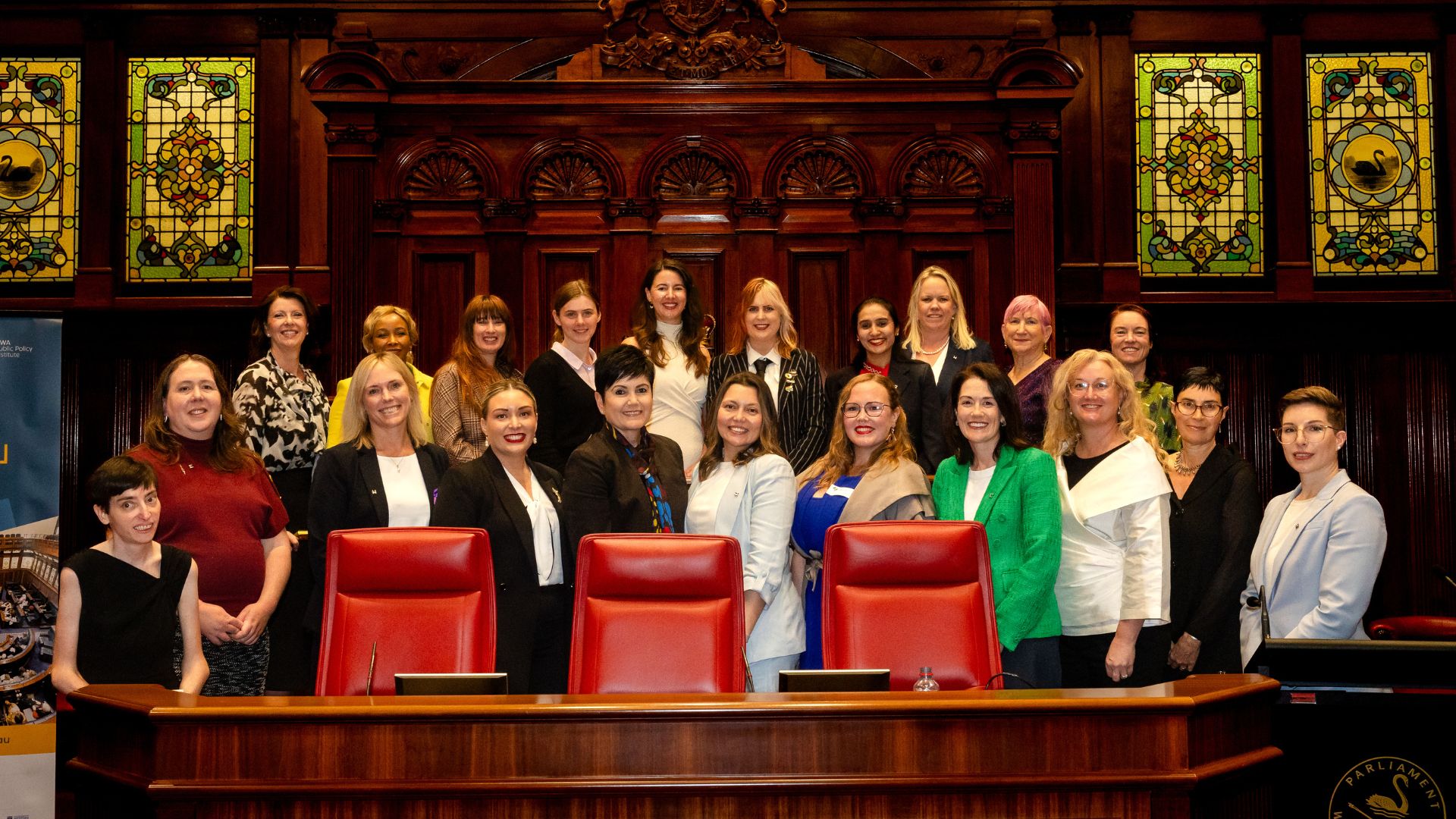 Group of 20+ smiling, diverse women, smartly dressed posing in a dark wood-panelled parliament chamber
