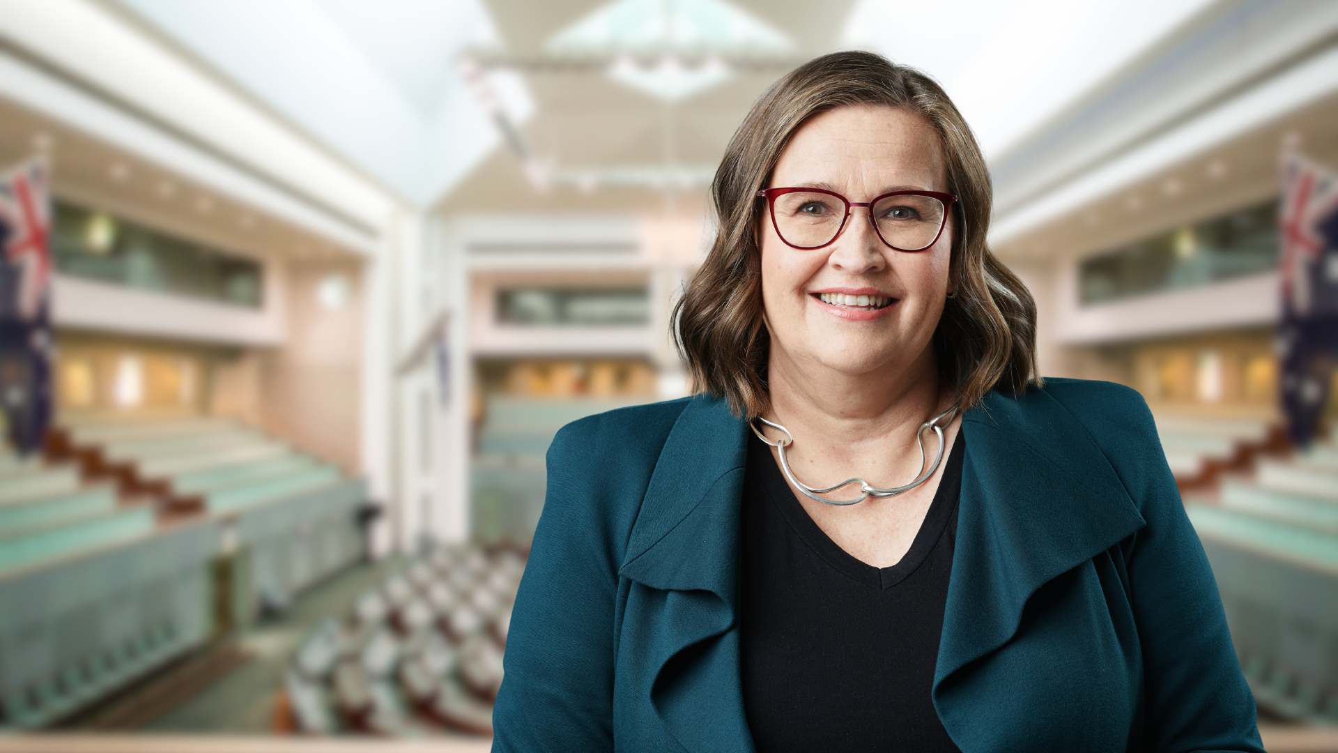 Photo of Kate Jenkins, a white woman with brown hair and glasses wearing a teal coloured jacket, against a blurred parliament chamber
