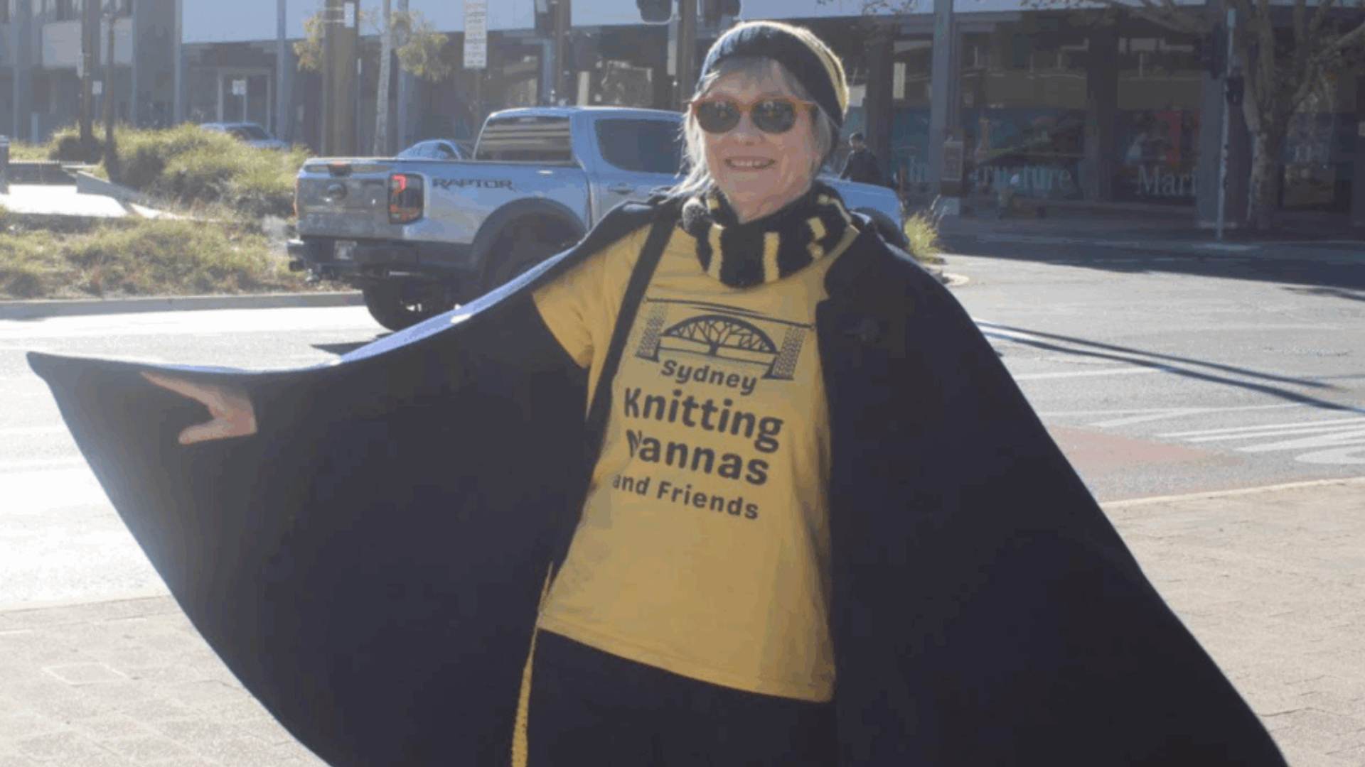 Photo of an older white oman wearing a black cape, sunglasses, and a yellow tshirt with the words "knitting nannas" printed on it