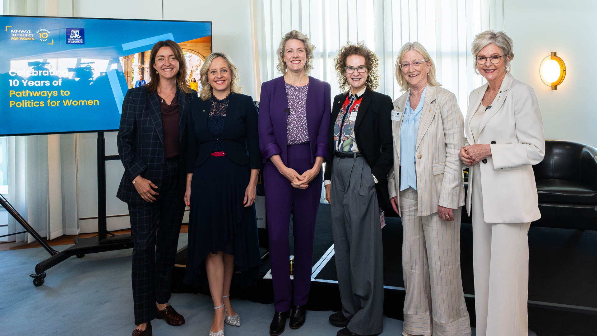 Six smartly dressed women pose for a photo standing in a line. A blue screen next to them reads "Celebrating 10 years of Pathways to Politics for Women"