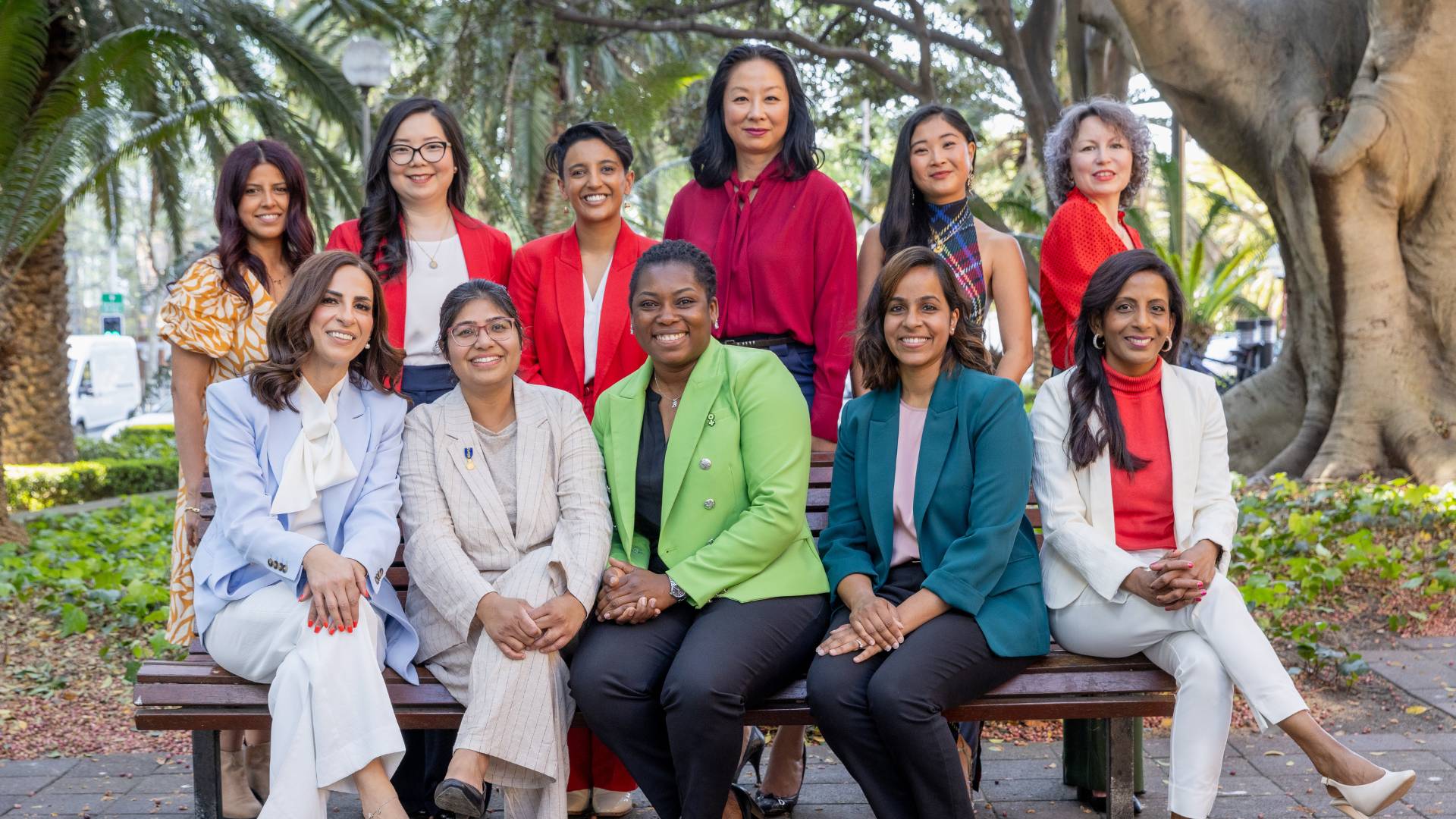 Diverse group of women pose for a photo sitting in two rows. They are wearing brightly coloured blazers and there are trees behind them.