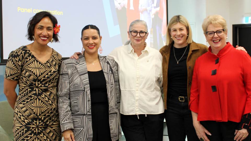 Five diverse women pose for a photo with their arms around each other, large projector screen in the background