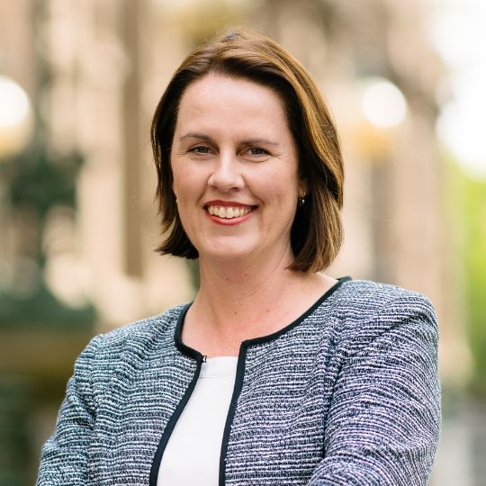 Smiling photo of Gaelle broad, brumette bobbed hair, wearing a casual blazer. Victorian Parliament building is out of focus behind her.