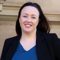 Profile photo of Pam Anderson, smiling, long straight black hair, wearing a black blazer, in front of an old stone building