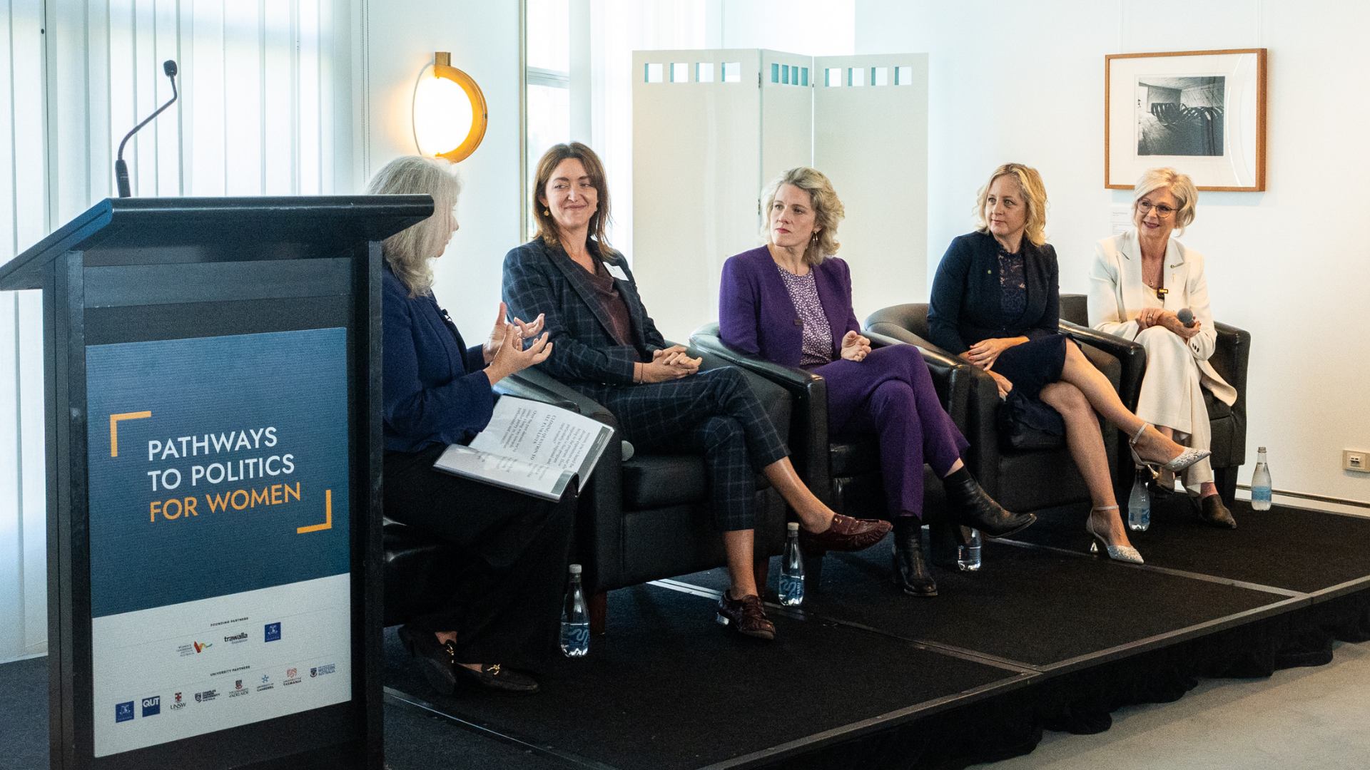 Five white women, smartly dressed, seated in a row on low stage
