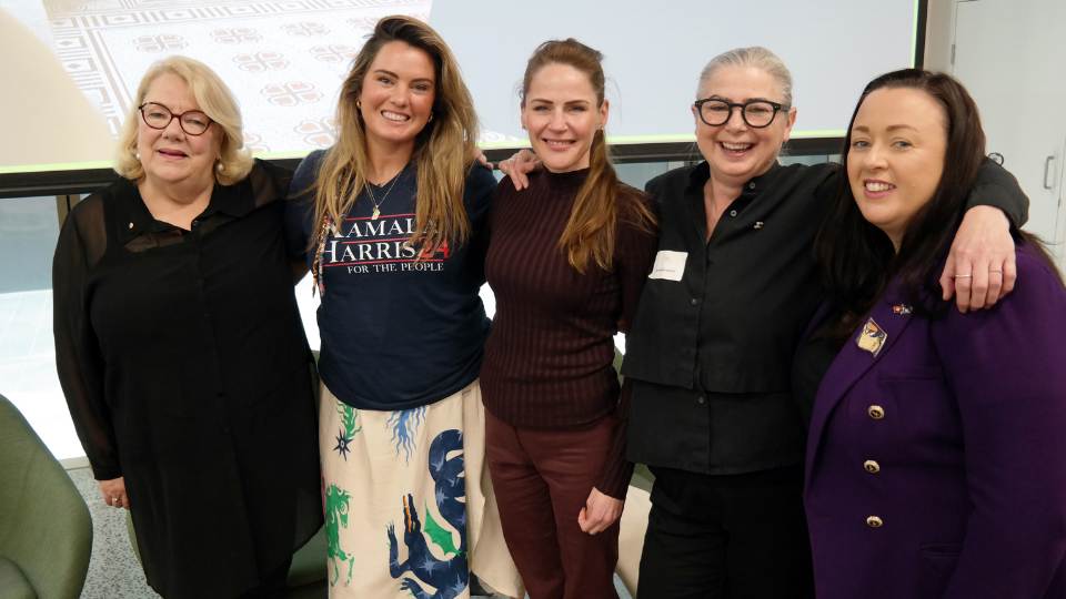 Group of five women, mostly dressed in black, smiling with their arms around each other, large projector screen in the background