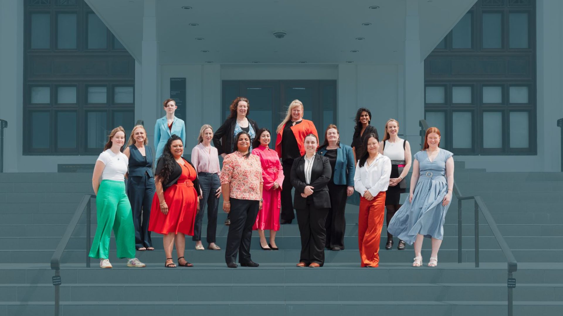 Photo of large group of women wearing colourful business dress, standing on the steps of a parliamentary building